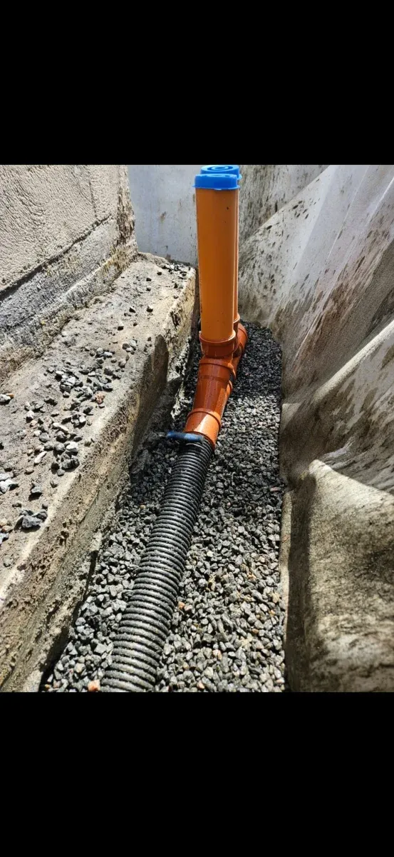 Svarta dräneringsrör i makadam längs husgrund med spolbrunn
